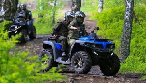 Two people on a blue ATV riding a trail with two more people riding an ATV behind them in a forest.