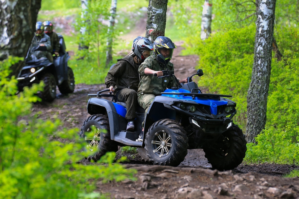 Two people on a blue ATV riding a trail with two more people riding an ATV behind them in a forest.