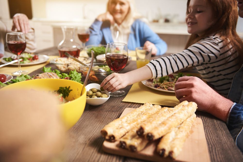 A girl reaches across a dinner table to grab a bowl of olives