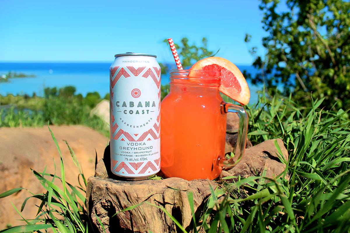 A can of Cabana Coast VOdka Greyhound and a mason jar cocktail sit in front of a lake