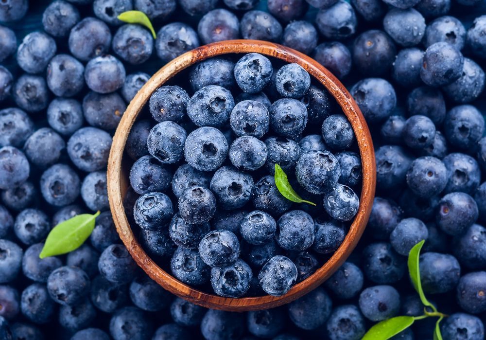A wooden bowl of blueberries surrounded by blueberries