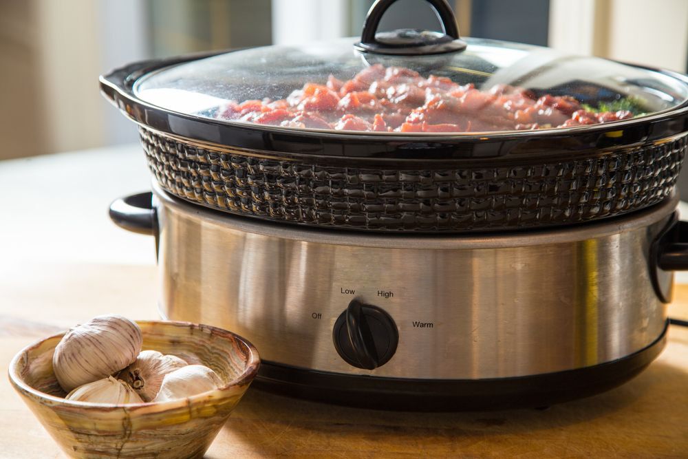 A photo of a crock pot on a table with a bowl of garlic beside it.