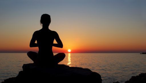 Silhouette of a person practicing meditation on a rock by the ocean at sunset.