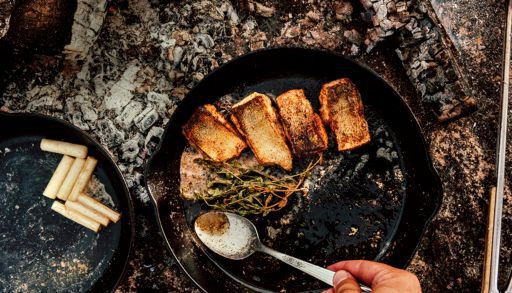 foraged pickerel roasting in a cast iron skillet over rocks while someone spoons butter overtop