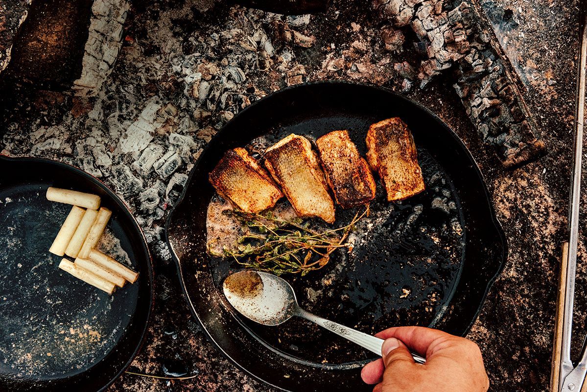 foraged pickerel roasting in a cast iron skillet over rocks while someone spoons butter overtop
