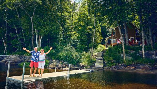 older retired couple waving goodbye from a dock in front of a cottage on a lake