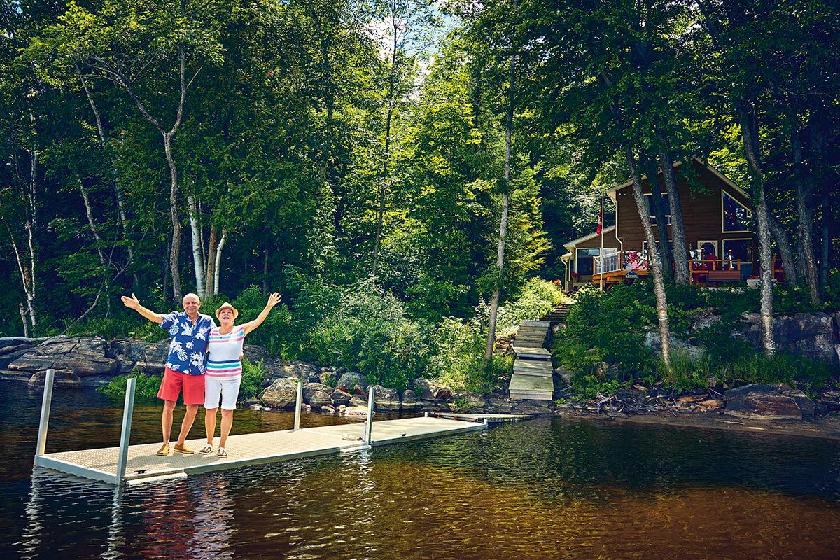 older retired couple waving goodbye from a dock in front of a cottage on a lake