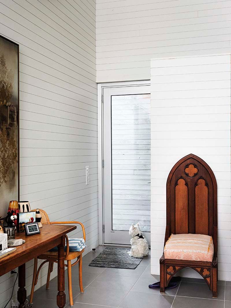 white interior with antique chair against one wall and a table against another near the front door. A small white dog looks out the door