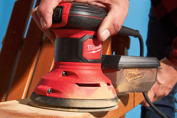 man using a random orbital sander to sand a muskoka chair on a blue background