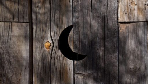 A vent in the shape of a half moon on an old distressed wooden outhouse door. There's knots and spaces in the wooden door. There's shadows on the wood.