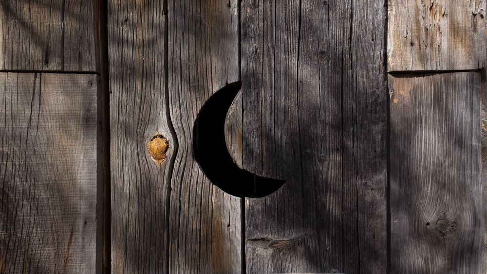 A vent in the shape of a half moon on an old distressed wooden outhouse door. There's knots and spaces in the wooden door. There's shadows on the wood.
