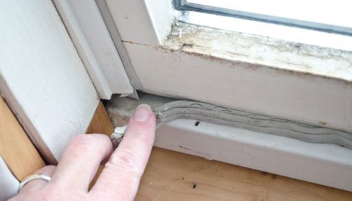 Caucasian woman's hand pressing in grey caulk in old window casement with bare wood windowsill for winterizing to keep out cold drafts and keep energy costs and heating bill down.