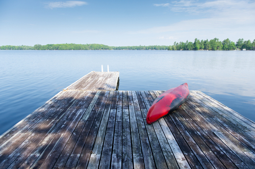 Old red kayak rest on a lake wooden dock on a summer day. Cottages are visible across the water.