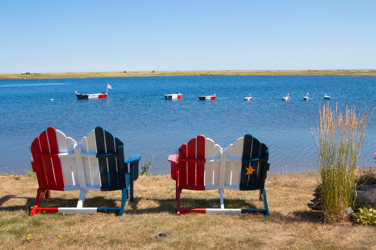 Wooden chairs along the shore and boat decorations on the water painted in the colors of the French Acadian flag on the coast near Bouctouche, New Brunswick.