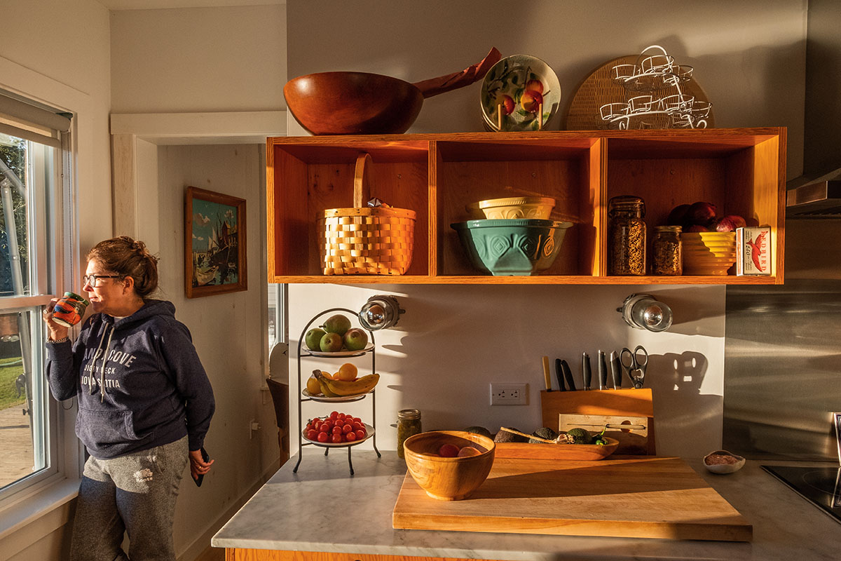 Pauline standing in her kitchen drinking a cup of coffee and looking out the window early in the morning