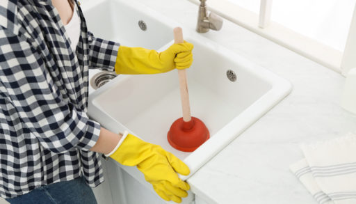 A woman with yellow rubber gloves holding a plunger in the kitchen sink