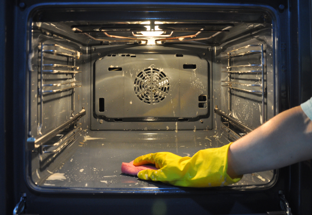 A yellow-gloved hand using a sponge to clean the inside of an oven