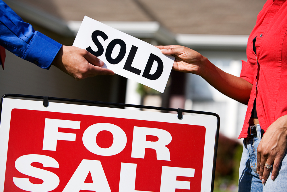 Two people exchanging a "SOLD" sign above a red-and-white For Sale sign