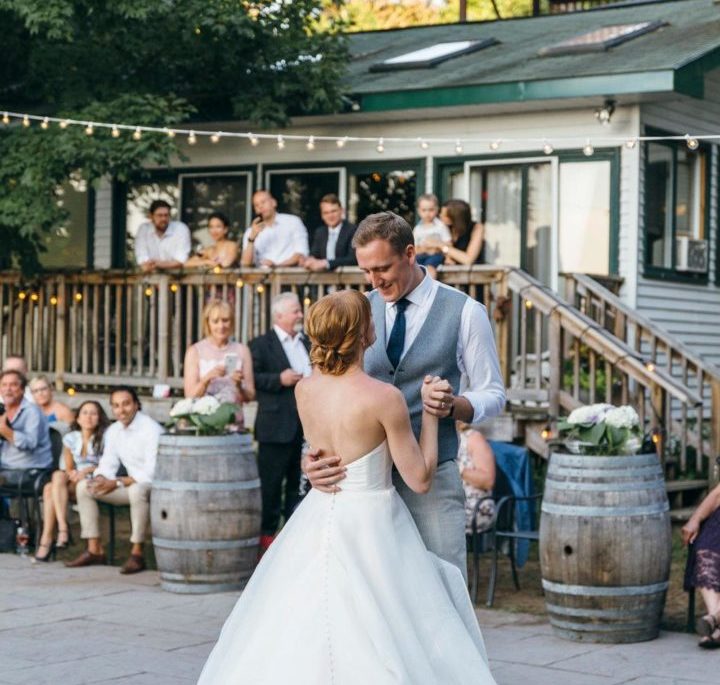 A couple dancing, dressed in wedding attire, outside in front of a cottage. Guests watching them in the background.