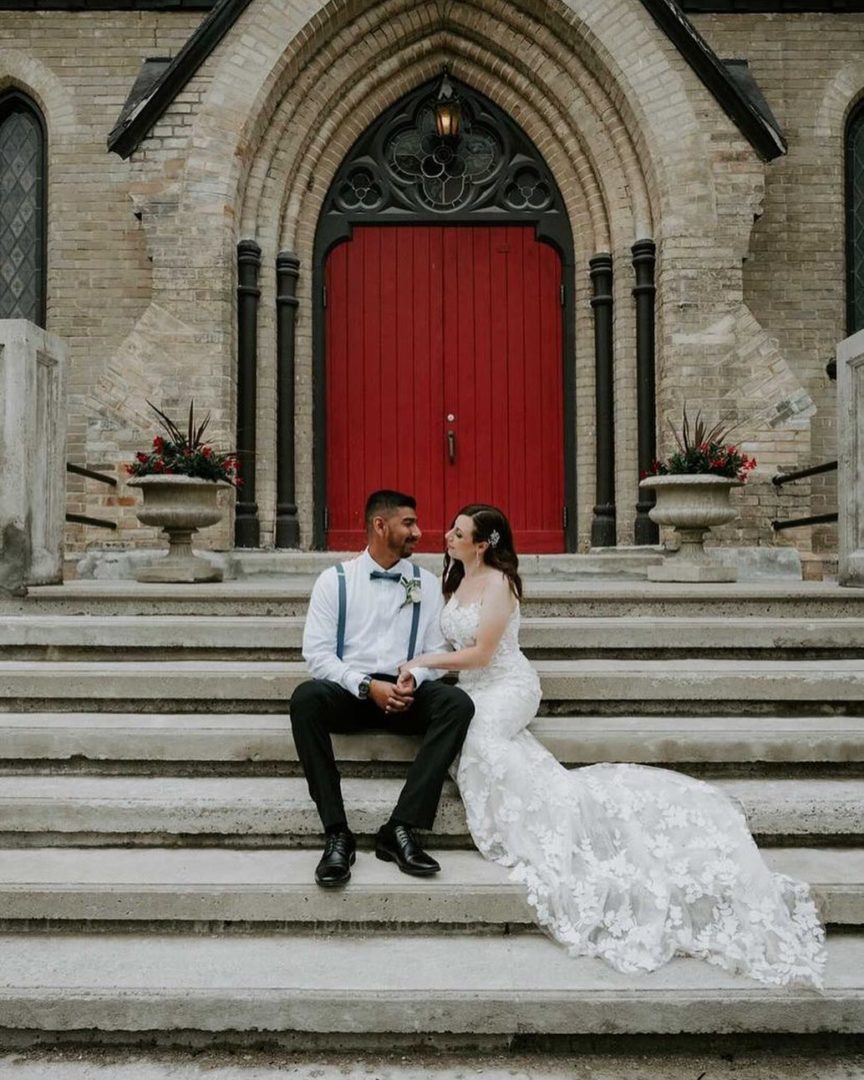 A couple sits in wedding attire on stone steps with a red door in the background.