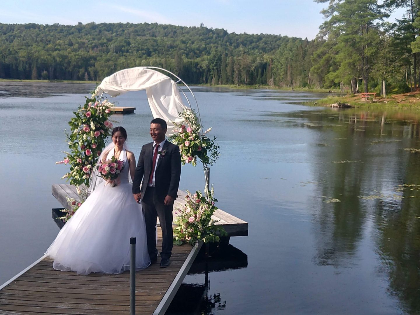 A couple stand in wedding attire on a dock in front of a large lake. A wedding arch with white chiffon and greenery is behind them.