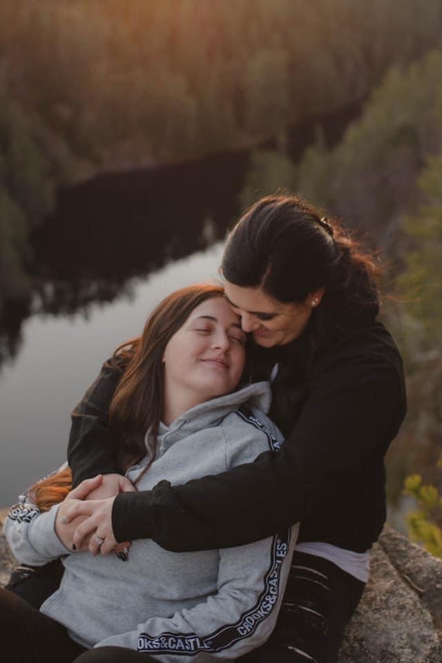 A couple embraces on a peak, with trees and a lake in the scenery below them. There is an engagement ring on one person's finger.