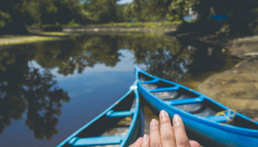 A person is holding out their hand with an engagement ring on, in front of a shoreline with blue canoes.