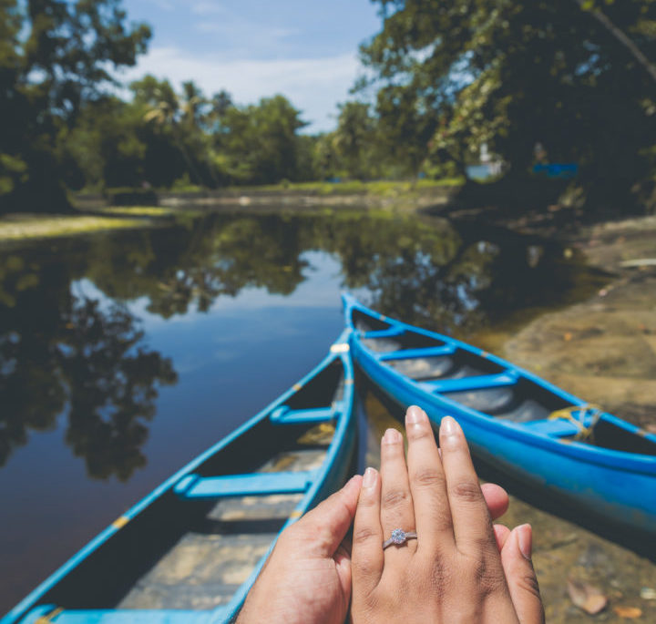 A person is holding out their hand with an engagement ring on, in front of a shoreline with blue canoes.