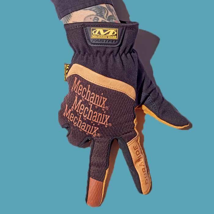 A hand wearing a black and orange Mechanix Wear Leather FastFit glove against a blue background.