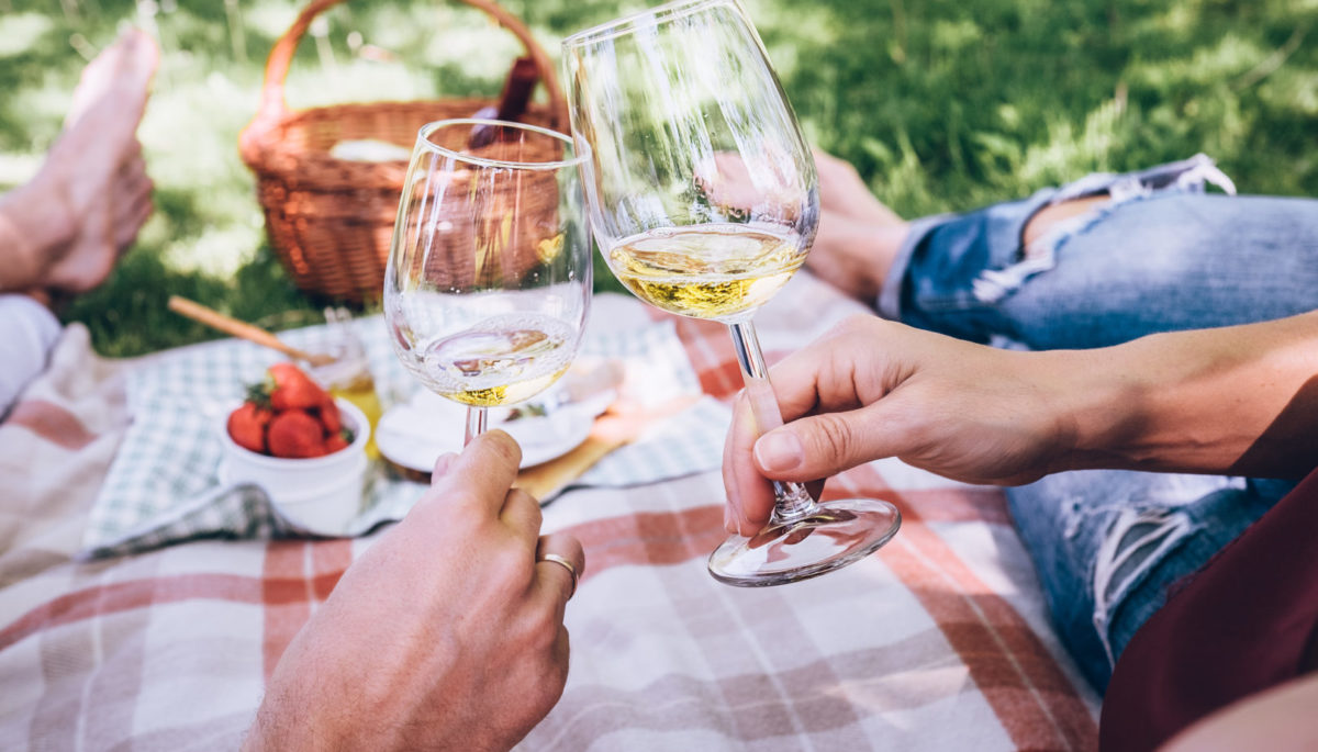 Two people on a summer date, clinking wine glasses on a checkered picnic blanket.