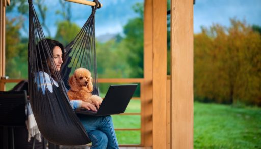 A woman on her deck working on a laptop, next to her dog.