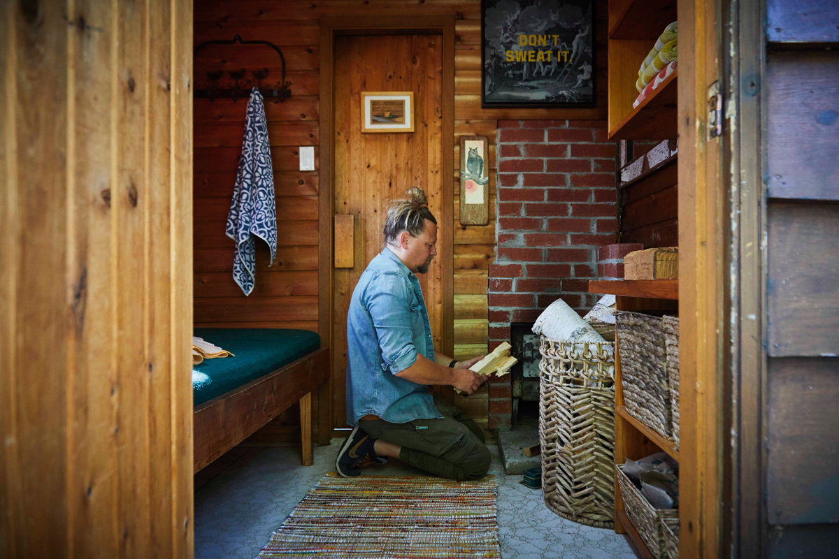 Mike Kelar sits on his knees in the Finnish sauna, feed wood into the stove