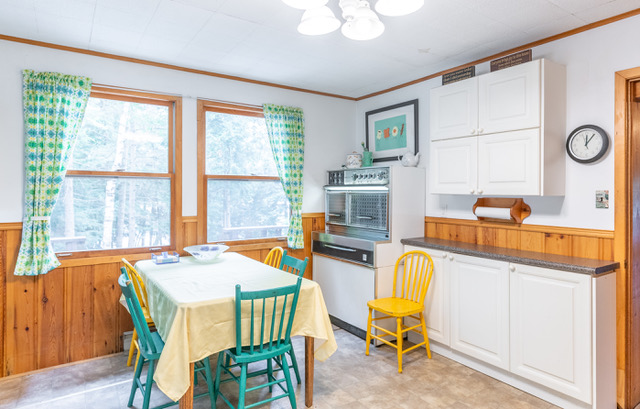 interior of a white kitchen with brown trim. An older stove sits in the corner with a table and multicoloured chairs