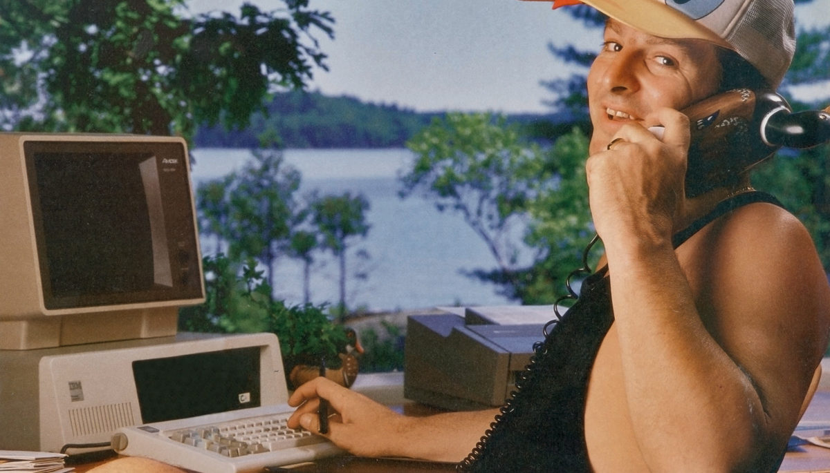 man in a donald duck hat sitting on a deck in cottage country holding an early cell phone in front of on an old computer
