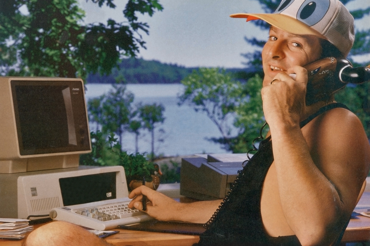 man in a donald duck hat sitting on a deck in cottage country holding an early cell phone in front of on an old computer