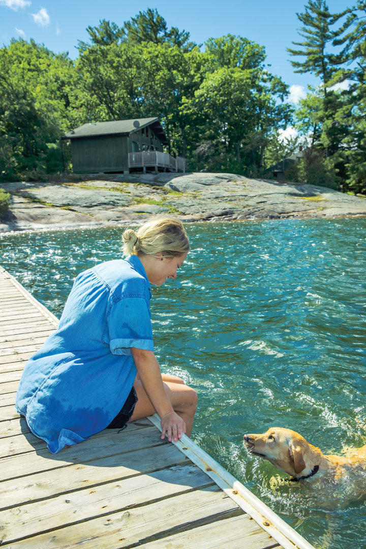 Lauren Patchett sits on the dock with her feet in the water as her dog swims