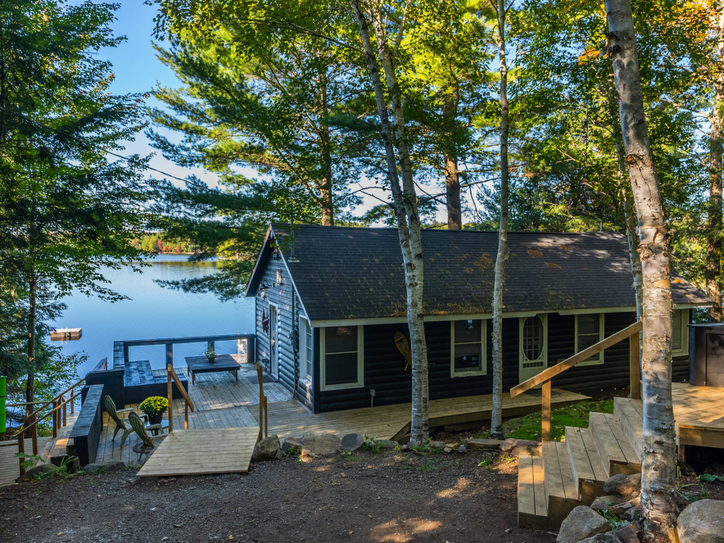 A cottage with a large deck, surrounded by tall trees with a lake in the background.