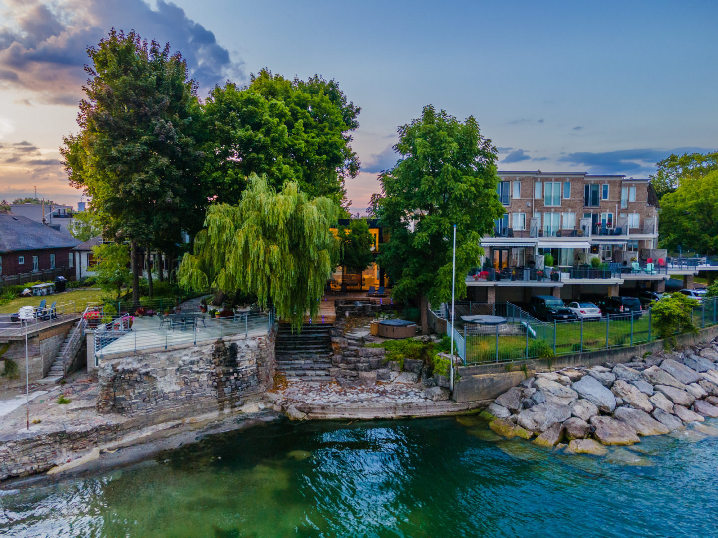 A small curve of waterfront with a modern home set back from the shoreline. Large windows in the house can be seen through a covering of trees, and there are neighbouring homes on either side of the house.