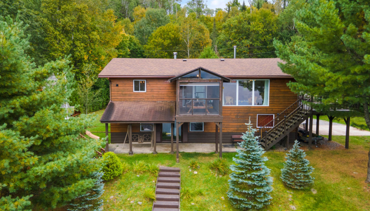 A large wood-panelled cottage sits on top of a hill, surrounded by trees with stairs heading down the slope.