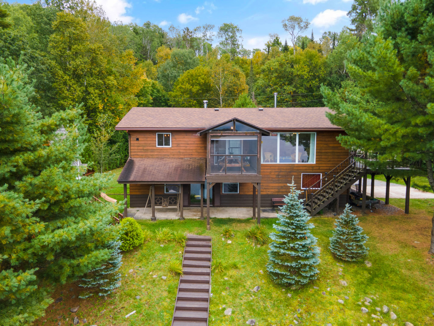 A large wood-panelled cottage sits on top of a hill, surrounded by trees with stairs heading down the slope.