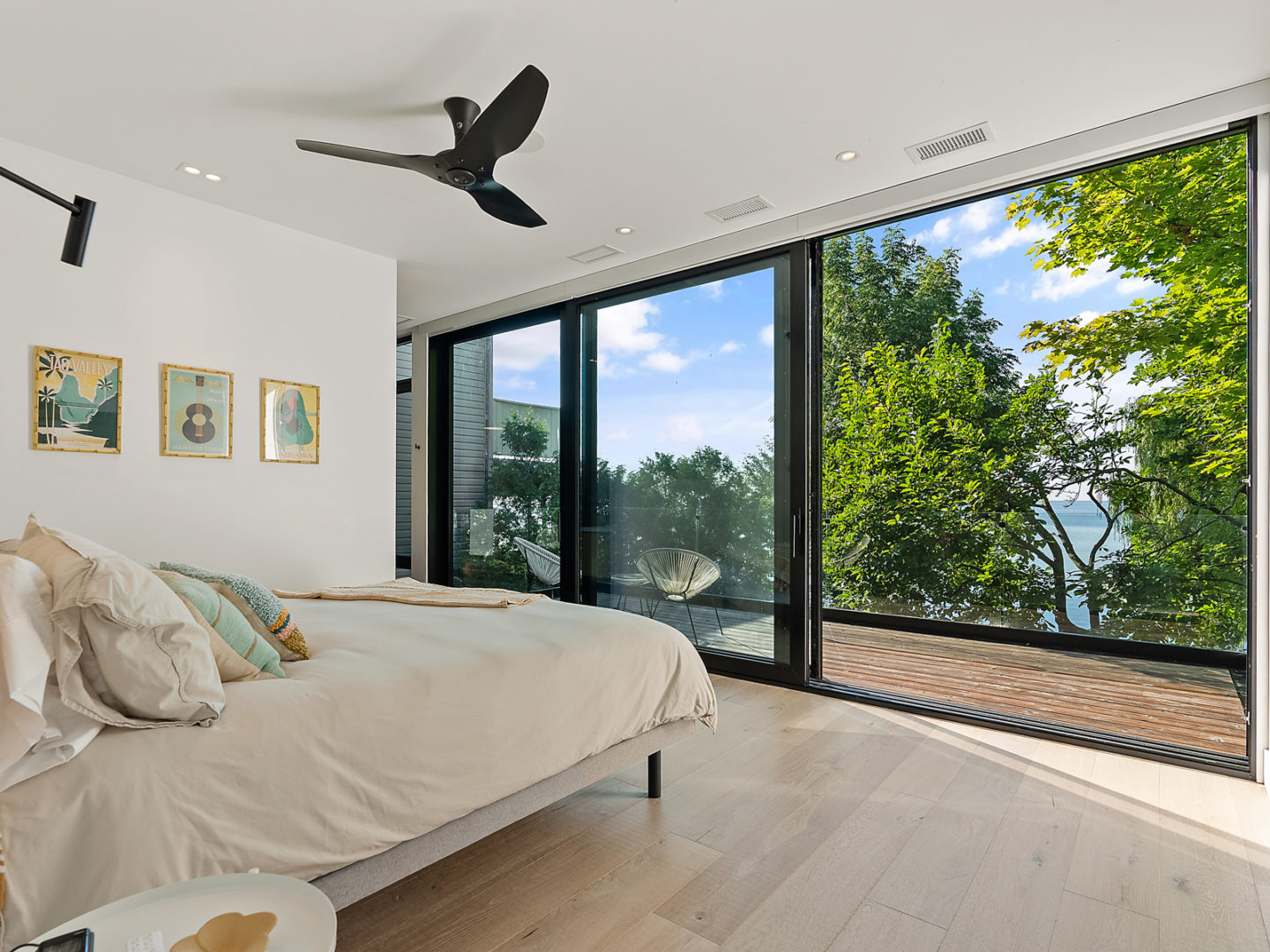 A large bedroom with light hardwood floors. A big bed looks out to a wall of floor-to-ceiling windows revealing a view of trees and lake.