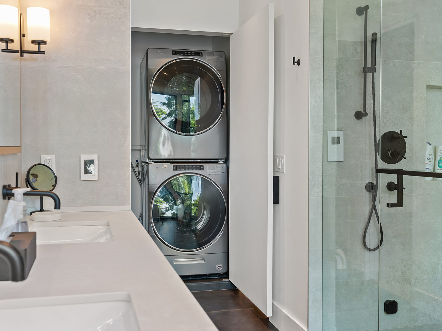 A bathroom area with a sink and a walk-in shower, with a stacked washing machine and dryer in a closet at the back of the room.