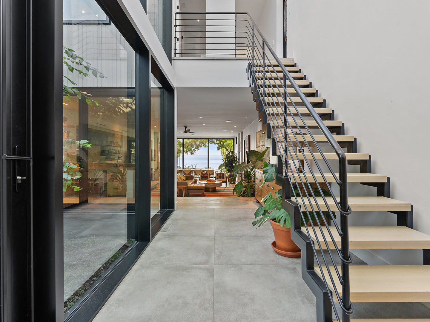 A long hallway that leads into an open-concept living area, with stairs leading up on the right side and a wall of windows on the other side.