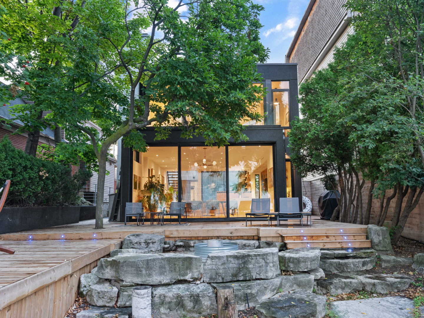 Exterior view of a modern, luxurious house with two storeys and floor-to-ceiling windows on both floors. A large deck extends outward, in front of the house.