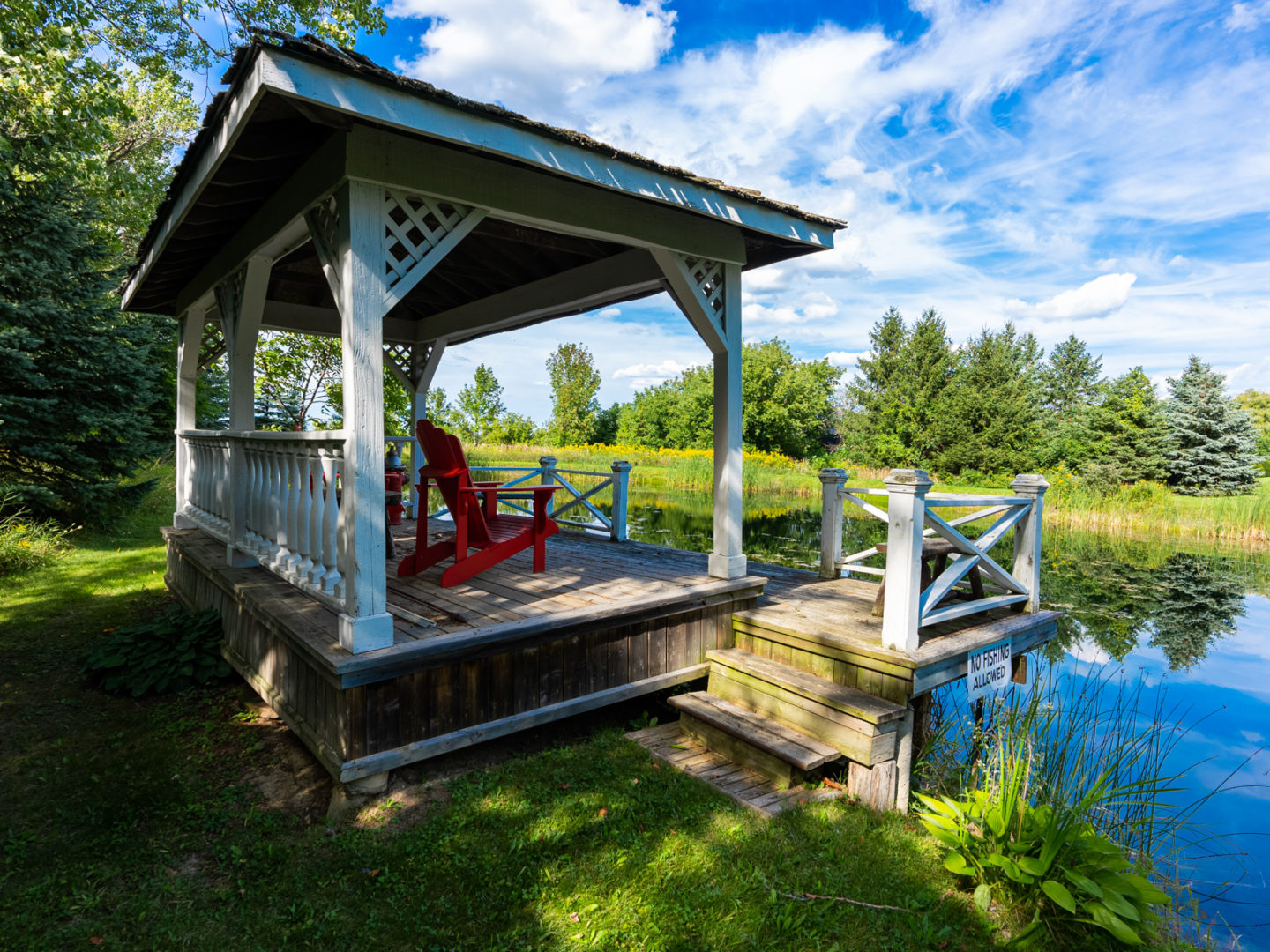 A covered wooden deck area with red Muskoka chairs, looking out over a small pond.
