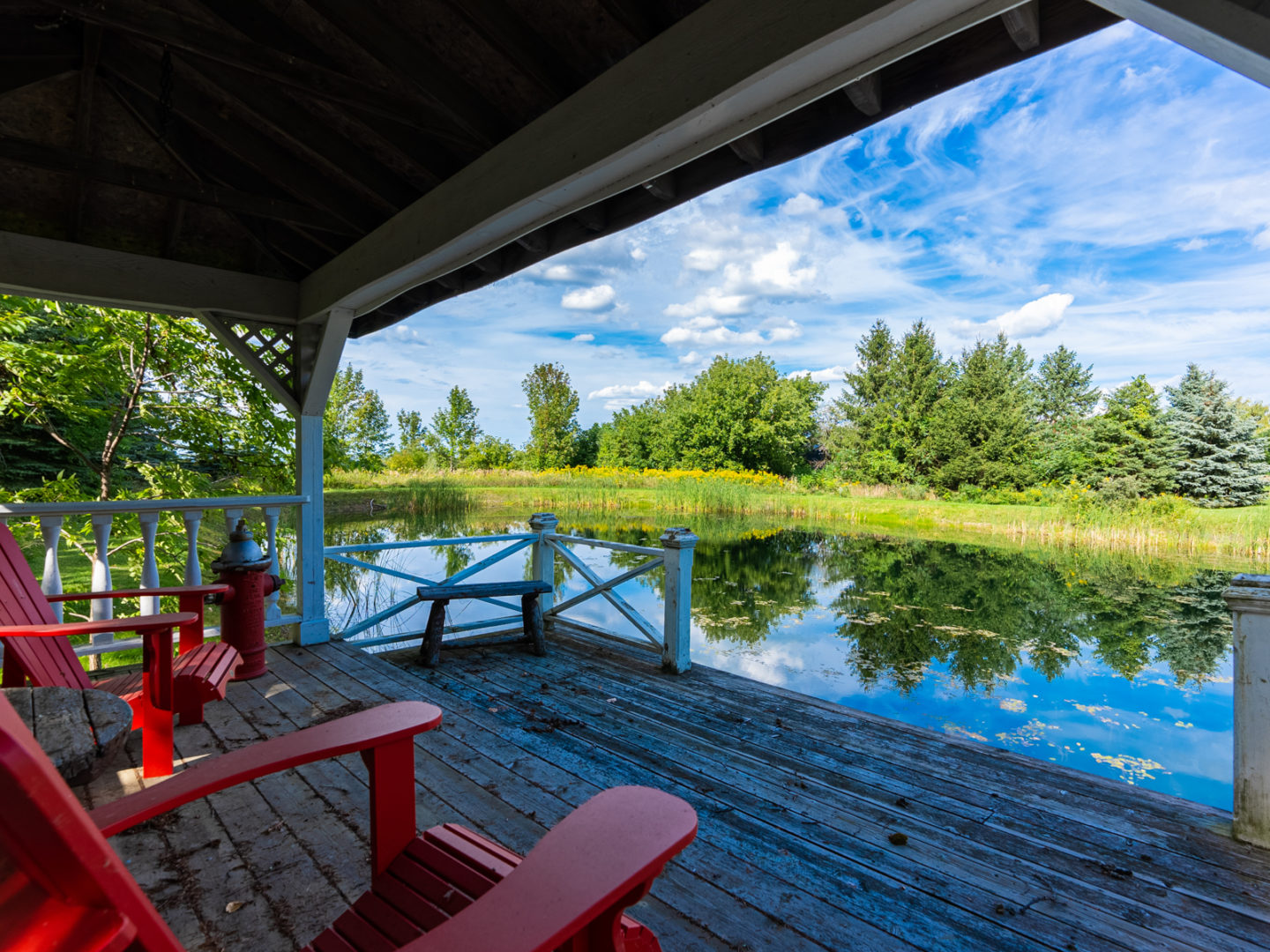 A covered wooden deck area with red Muskoka chairs, looking out over a small pond.
