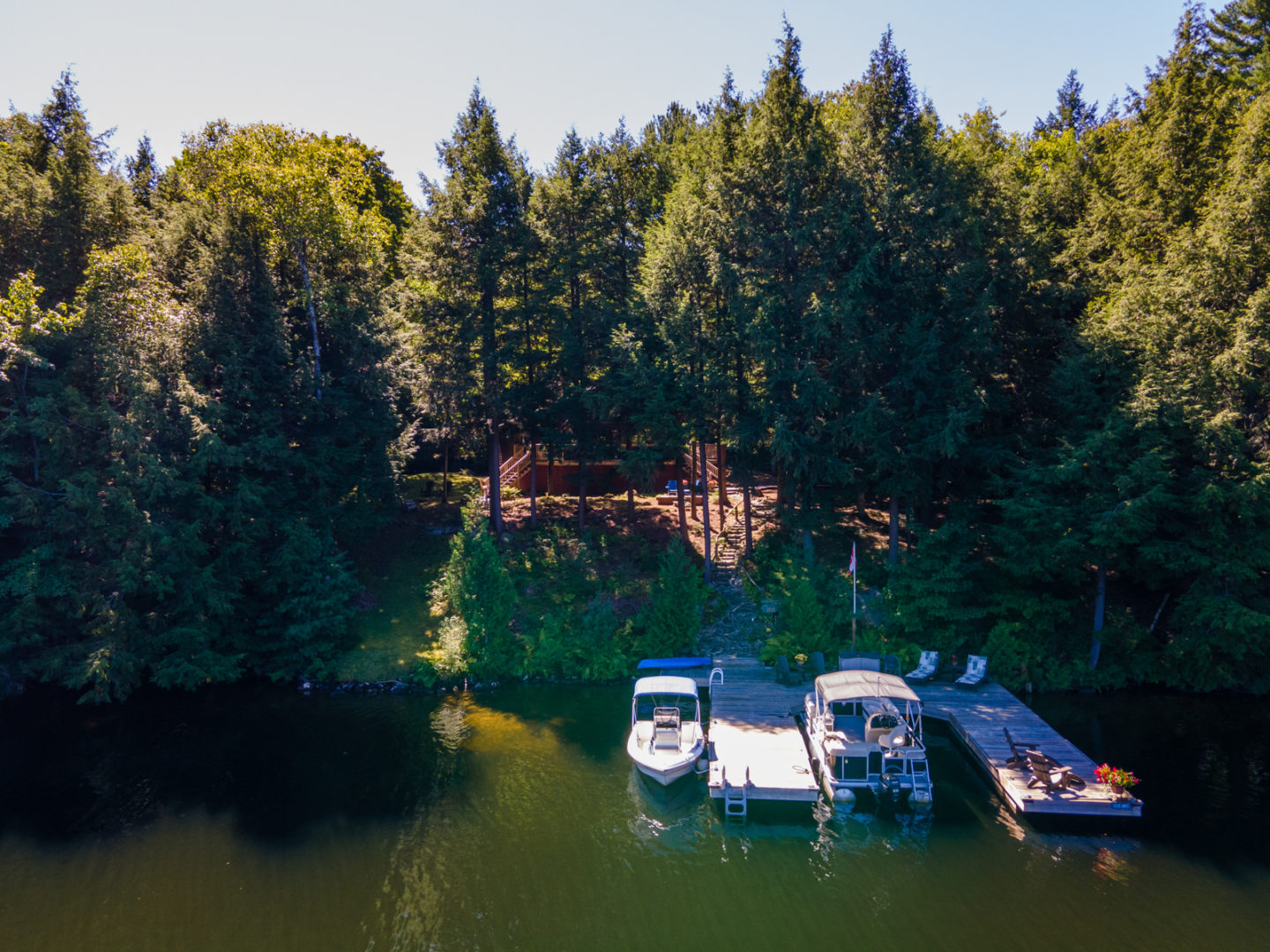 Waterfront view of a cottage on a sloping waterfront, surrounded by trees. A large dock extends out front into the lake.