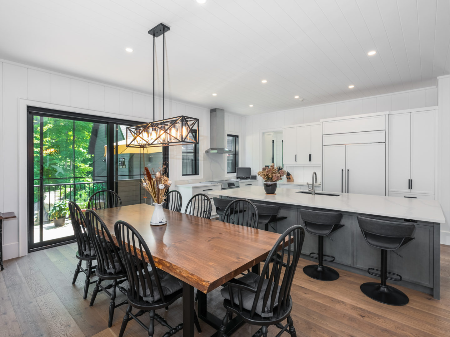 An open-concept kitchen and dining space with a big table beside a large island. White countertops, white cupboards, and large windows add brightness.