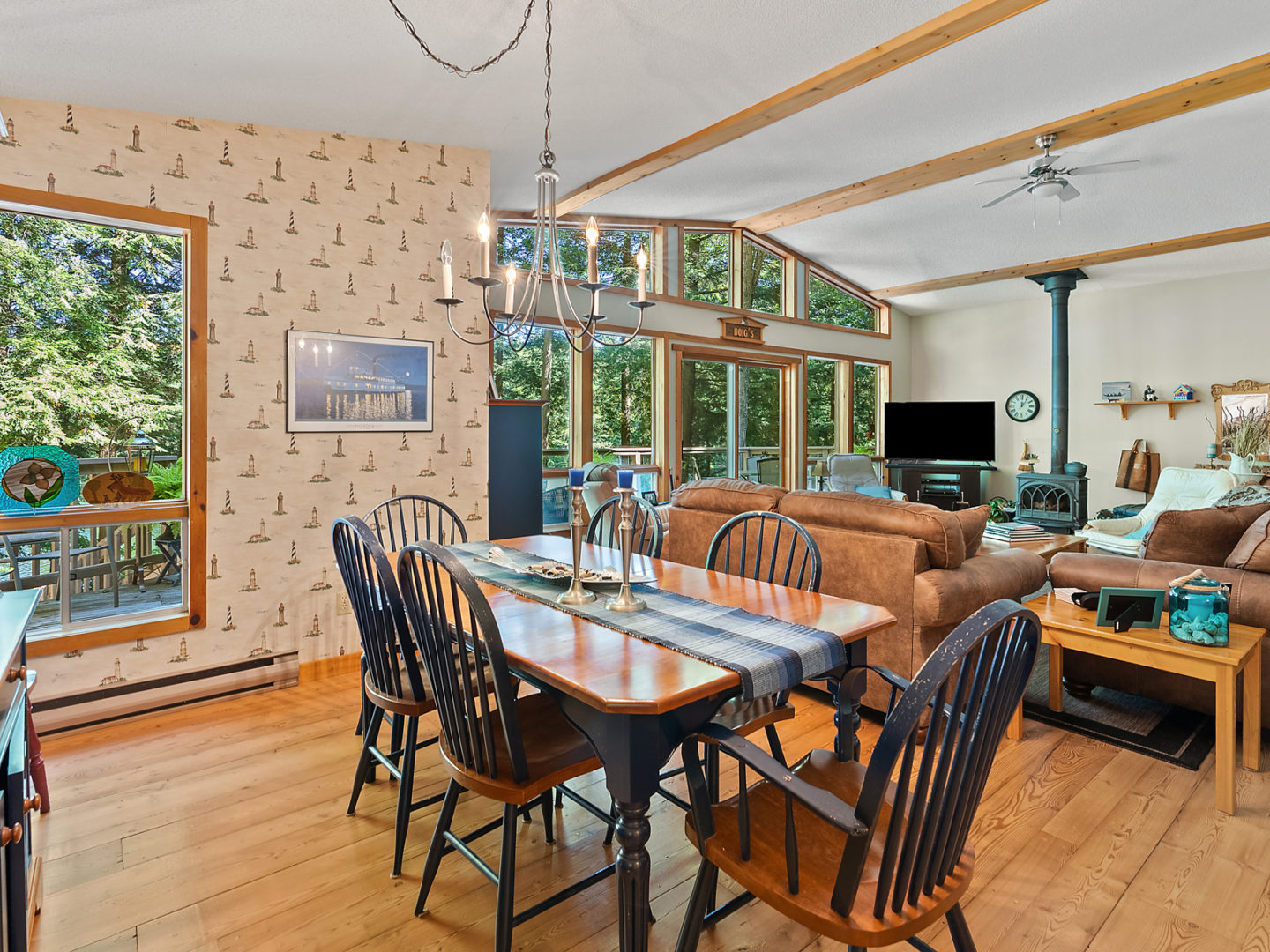An open-concept cottage living space. A small dining area with a table and chairs transitions into a living room with couches, a fireplace, and floor-to-ceiling windows across one wall.