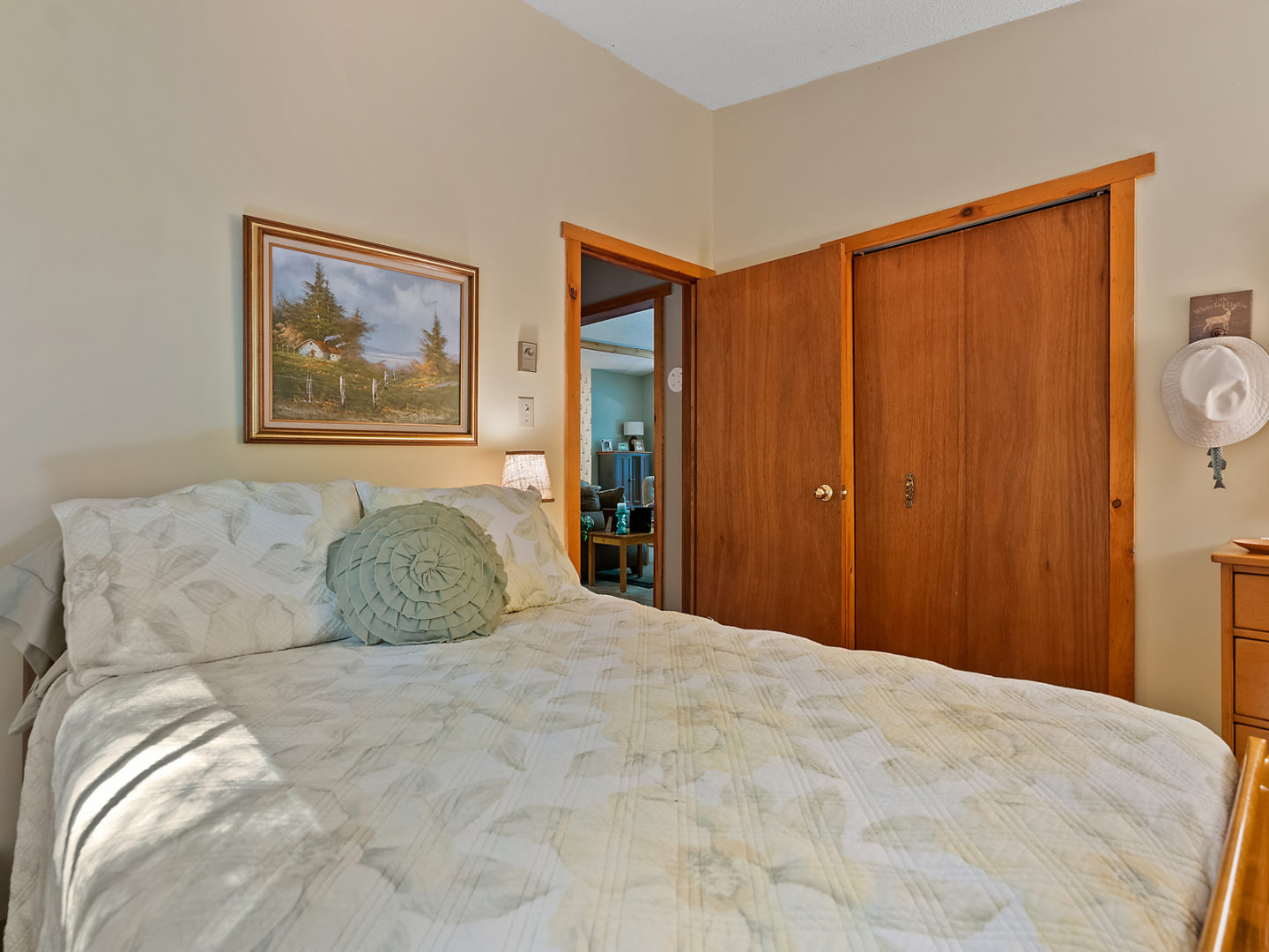 A small bedroom with a bed and a closet. An open door looks out to a living room.